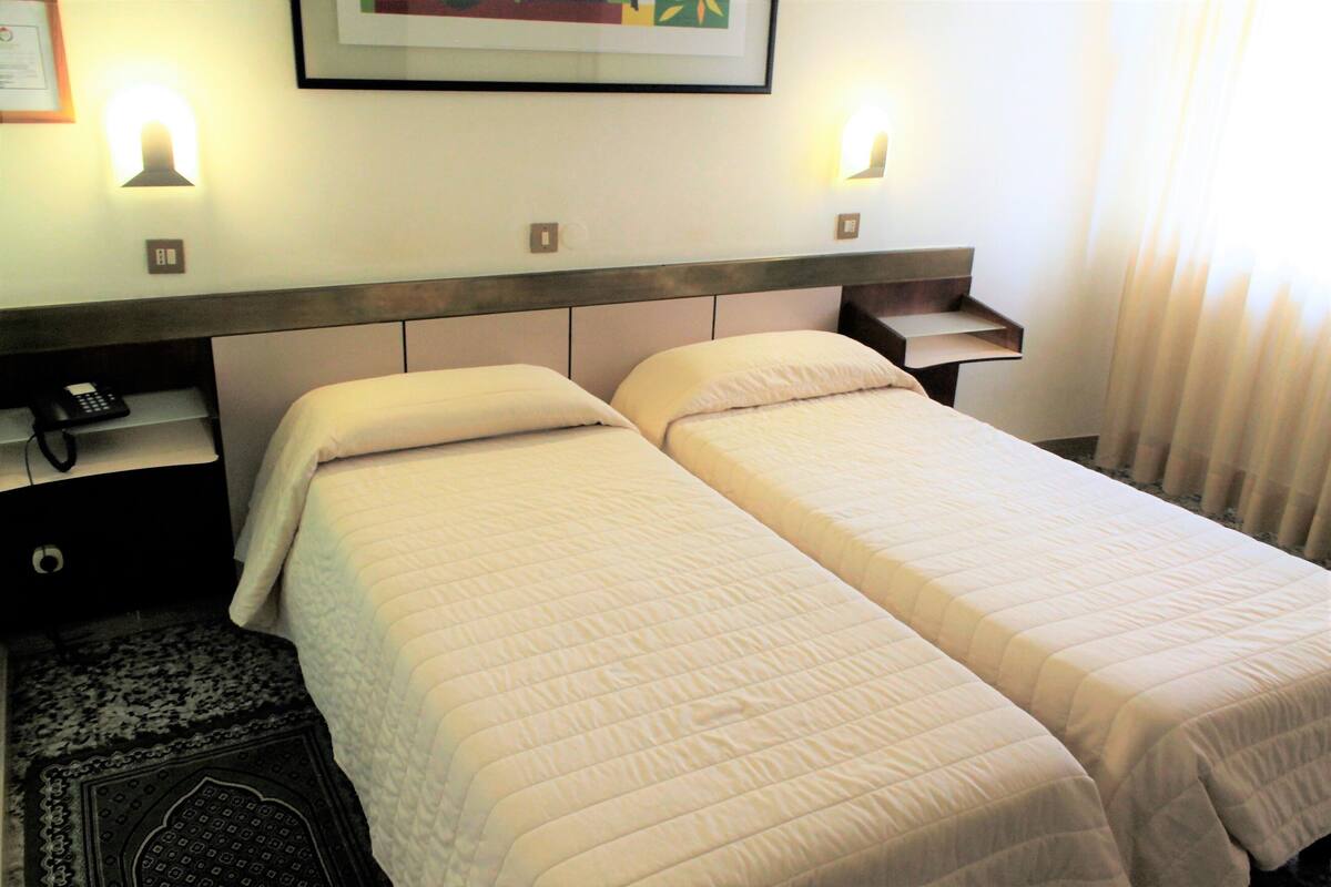A hotel room with two single beds, a nightstand with a phone, and a wall-mounted light fixture.