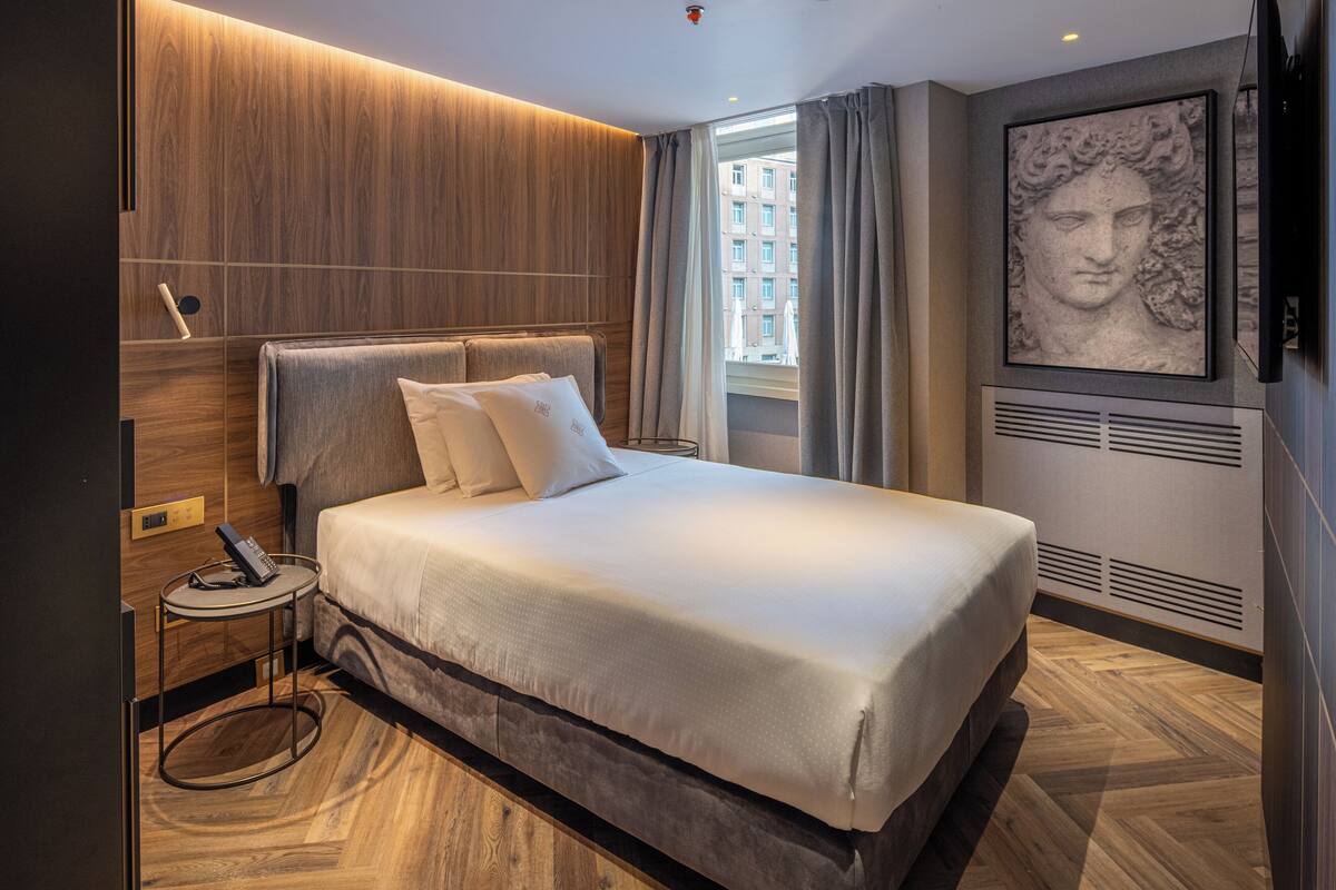 Camera d'albergo moderna con un letto grande, un comodino con telecomando, una TV a parete e un'ampia finestra con tende.