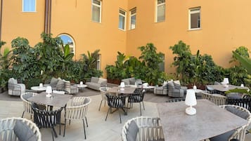 Area esterna con diversi tavoli e sedie, immersa nel verde e con un edificio con ampie finestre.