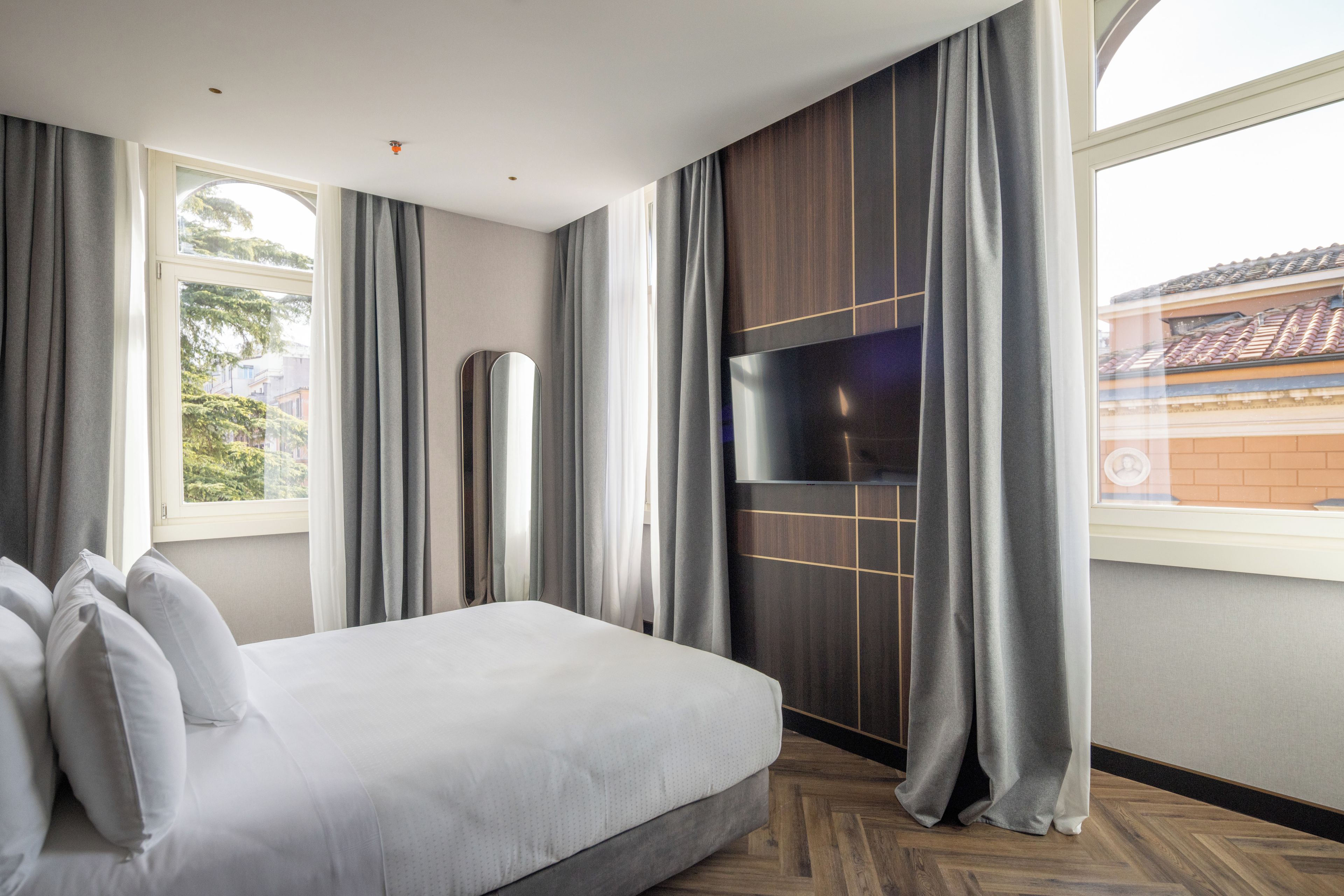 A modern hotel room with a large bed, a flat-screen TV, and a view of the city through a window.