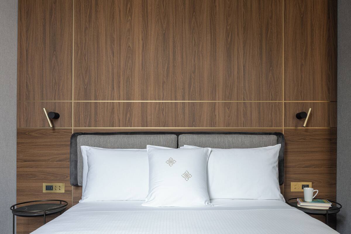 Una camera d'hotel con una testiera in legno, un letto con biancheria da letto bianca, due comodini con lampade e un libro su uno dei tavolini.