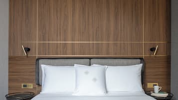 Un letto rifatto con cura, lenzuola bianche e due cuscini, un comodino con un libro e una tazza, e una testiera in legno dal design moderno.