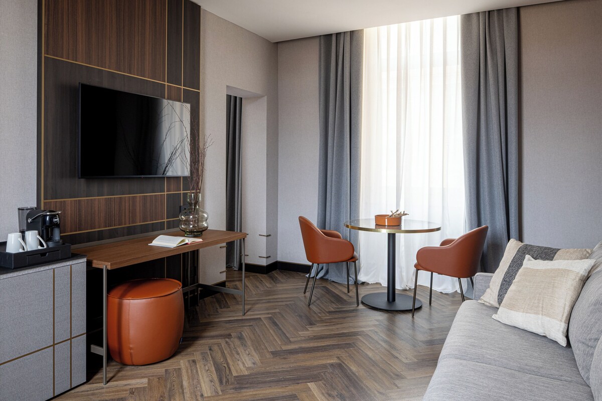A modern hotel room with a flat-screen TV, a wooden desk, a round dining table, two orange chairs, a grey sofa, and a large window with curtains.