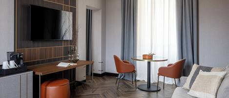 A modern hotel room with a flat-screen TV, a wooden desk, a round dining table, two orange chairs, a grey sofa, and a large window with curtains.
