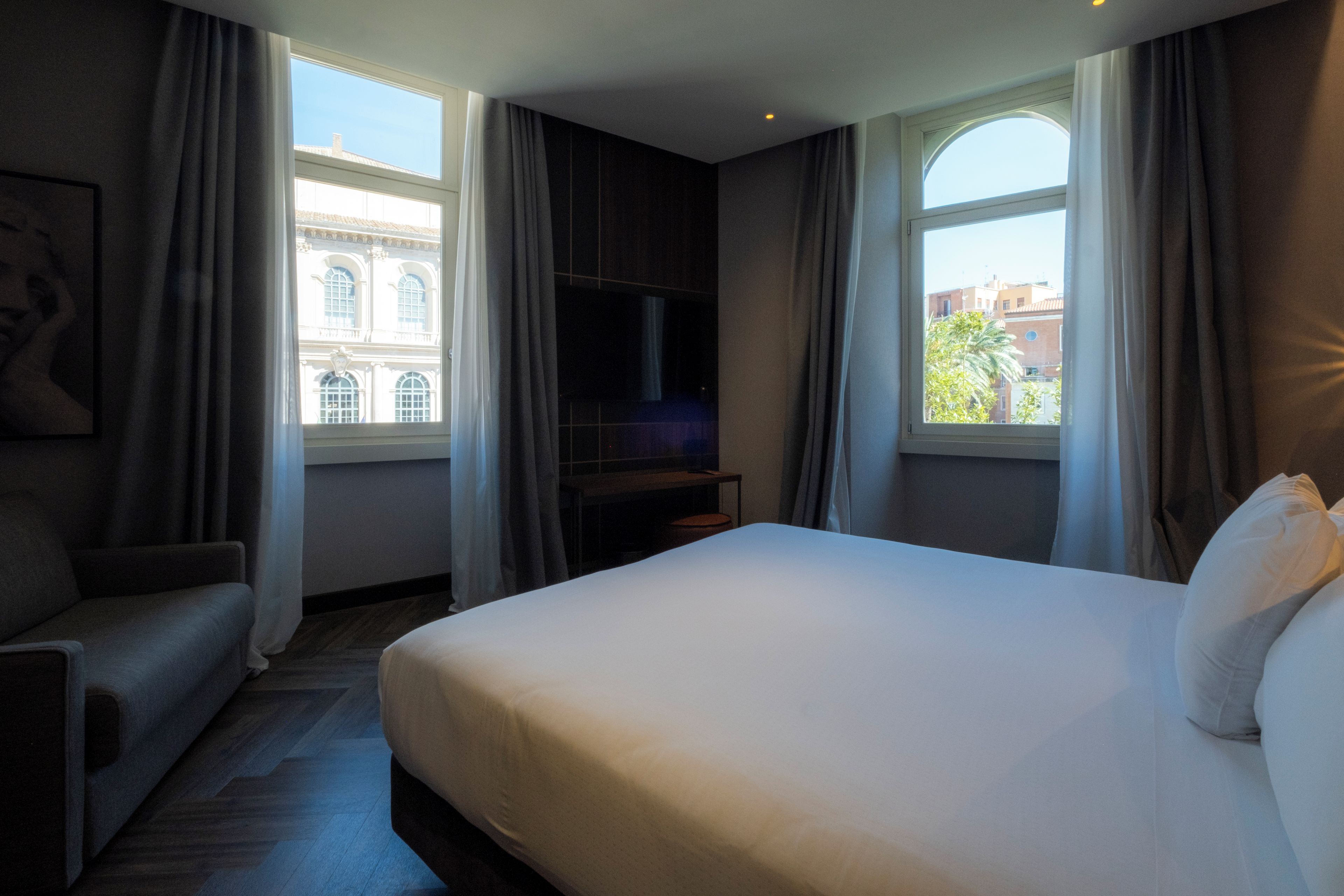 Una habitación de hotel con cama, televisión, sofá y ventanas grandes con cortinas.