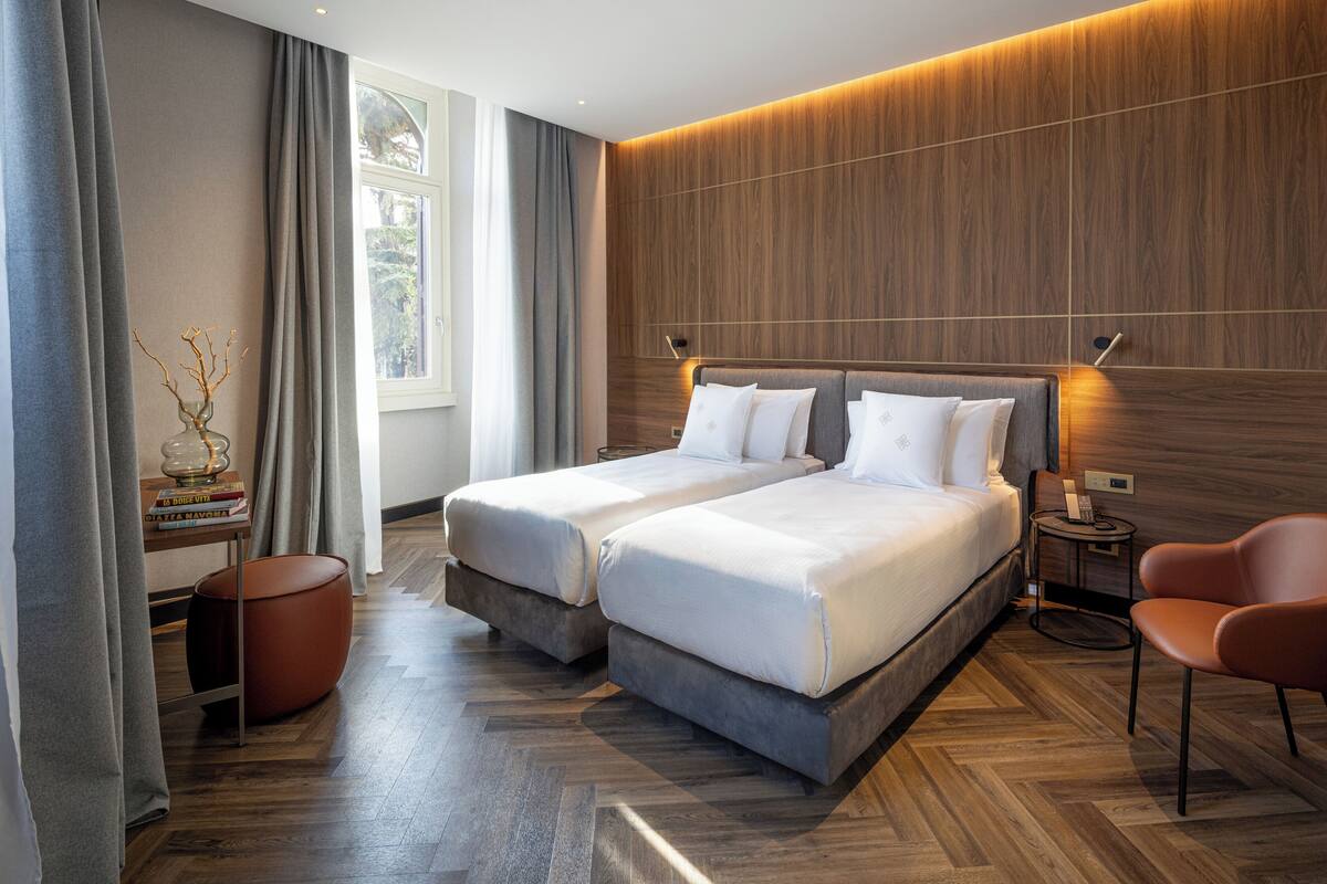 Camera d'albergo moderna con due letti, una parete decorata in legno, un comodino, una sedia e una finestra con tende.