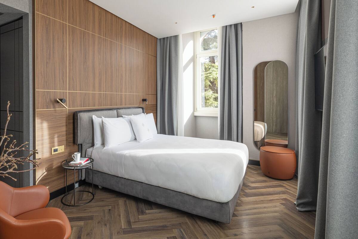 Una moderna camera d'hotel con un letto grande, un tavolino, un pouf arancione rotondo, uno specchio ampio e vista su un'area verde.