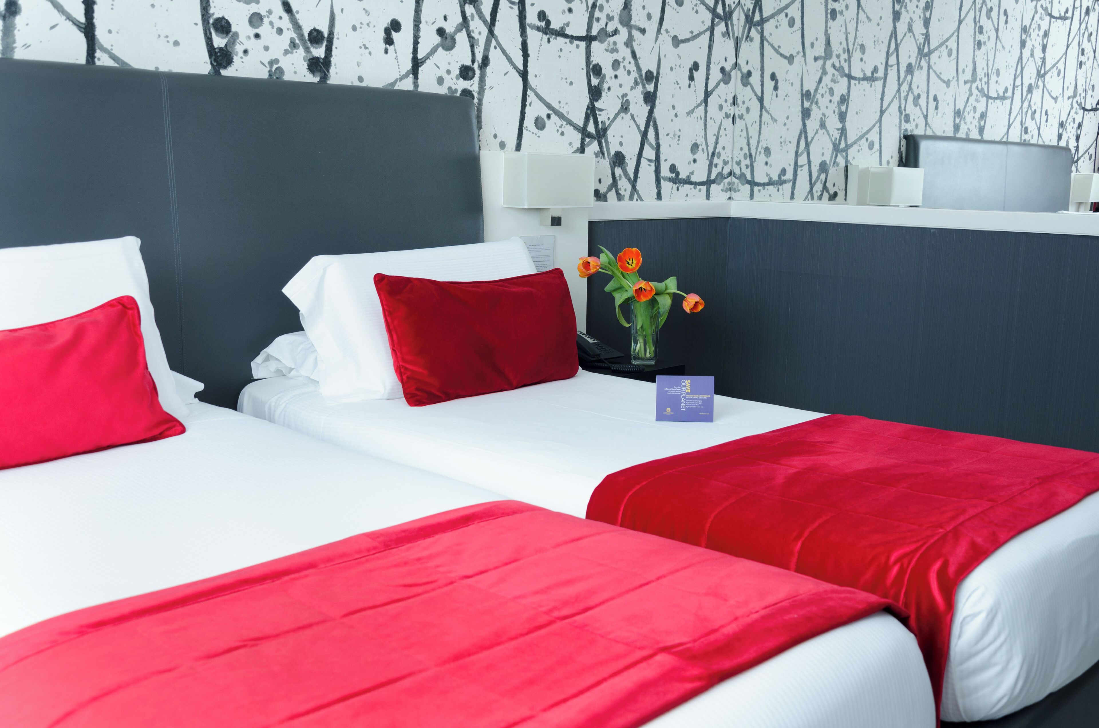 Habitación de hotel con dos camas, cada una con ropa de cama blanca y detalles rojos, un jarrón con flores naranjas y un diseño de pared moderno.