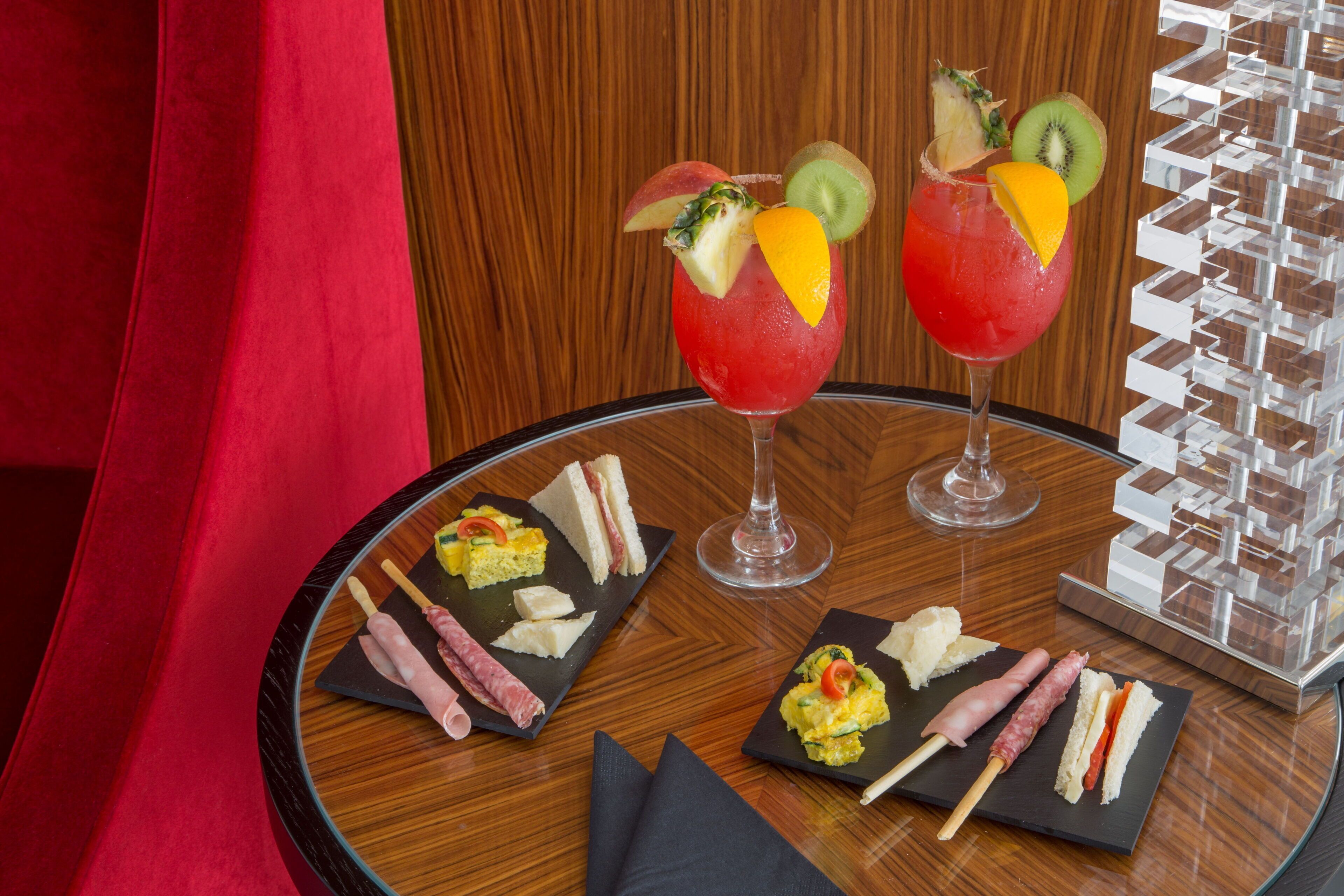 Una mesa redonda con superficie de vidrio, dos bebidas adornadas con fruta y diferentes alimentos en platos negros.