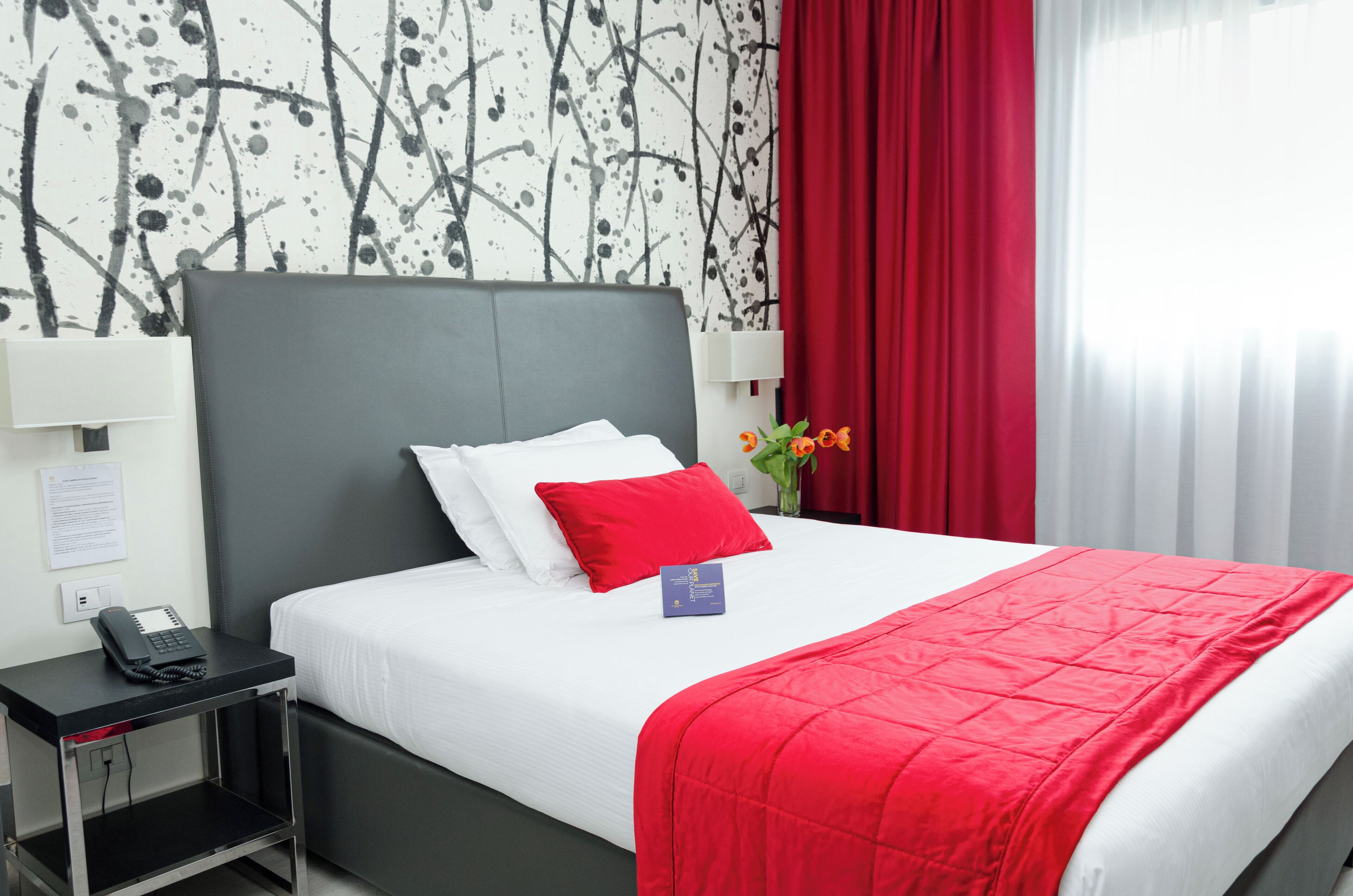 Een hotelkamer met een bed, een nachtkastje met een telefoon, een rood gordijn en een muur met een zwart-wit abstract ontwerp.