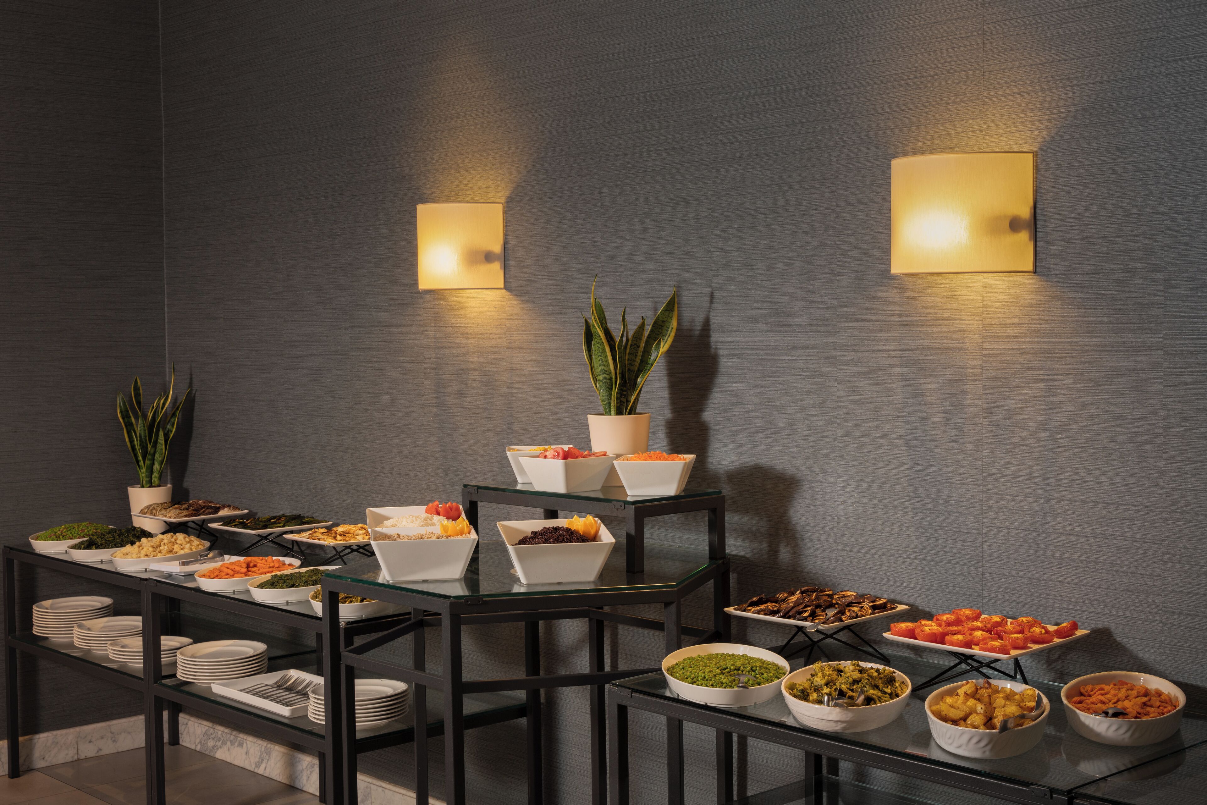 A buffet setup with various food items on glass tables, surrounded by black chairs, against a textured wall with two wall-mounted lights.