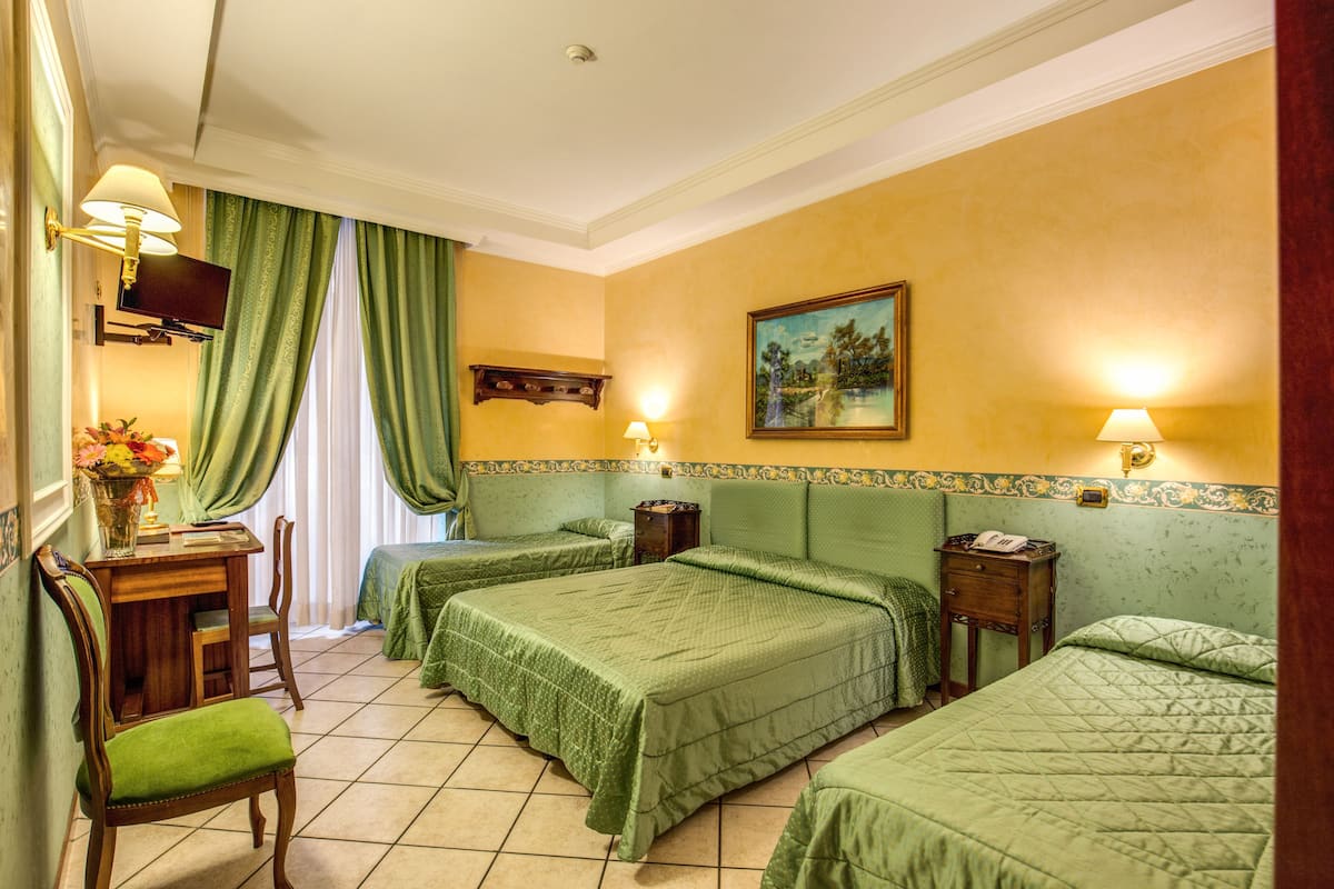 A hotel room with two beds, a desk, and a chair. There is a painting on the wall and a lamp on each side of the bed.
