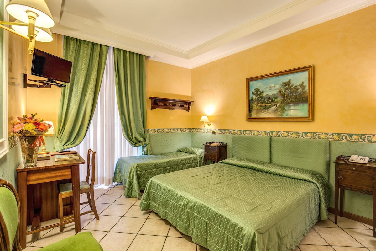 A hotel room with two beds, a desk, a chair, a TV, a painting, and a lamp.