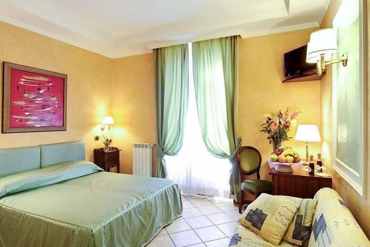 A hotel room with a bed, a sofa, a table with a lamp, a wall-mounted TV, and a window with curtains.
