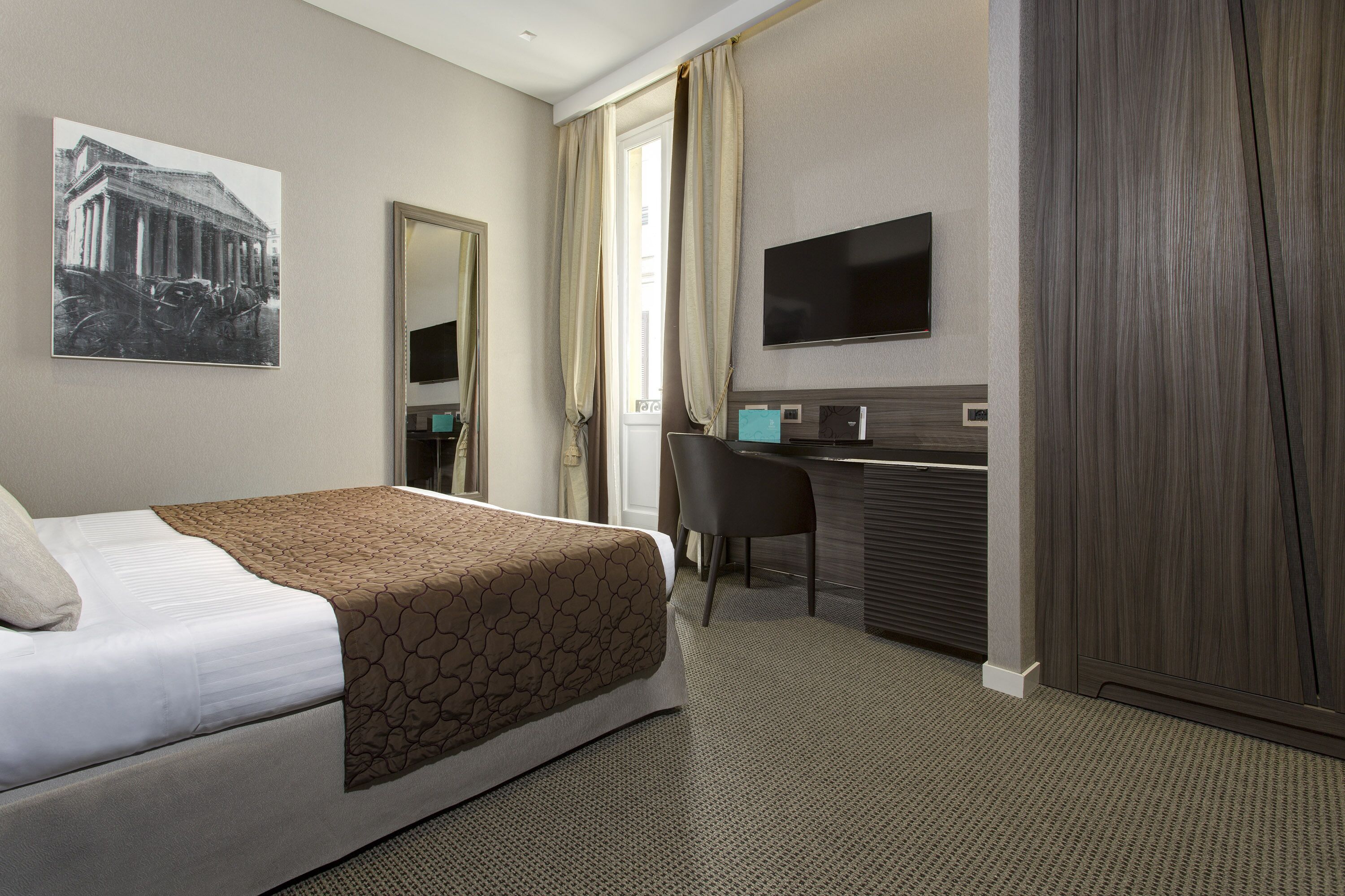 A hotel room with a bed, a desk, a television, and a large window with curtains.