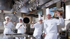 A professional kitchen with stainless steel appliances, a large pot on the counter, and chefs in white uniforms. - Hotel Artemide (Rome)