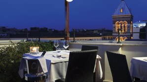 An outdoor dining area with a table set for two, a candle, and a view of a building with a tower. - Hotel Artemide (Rome)