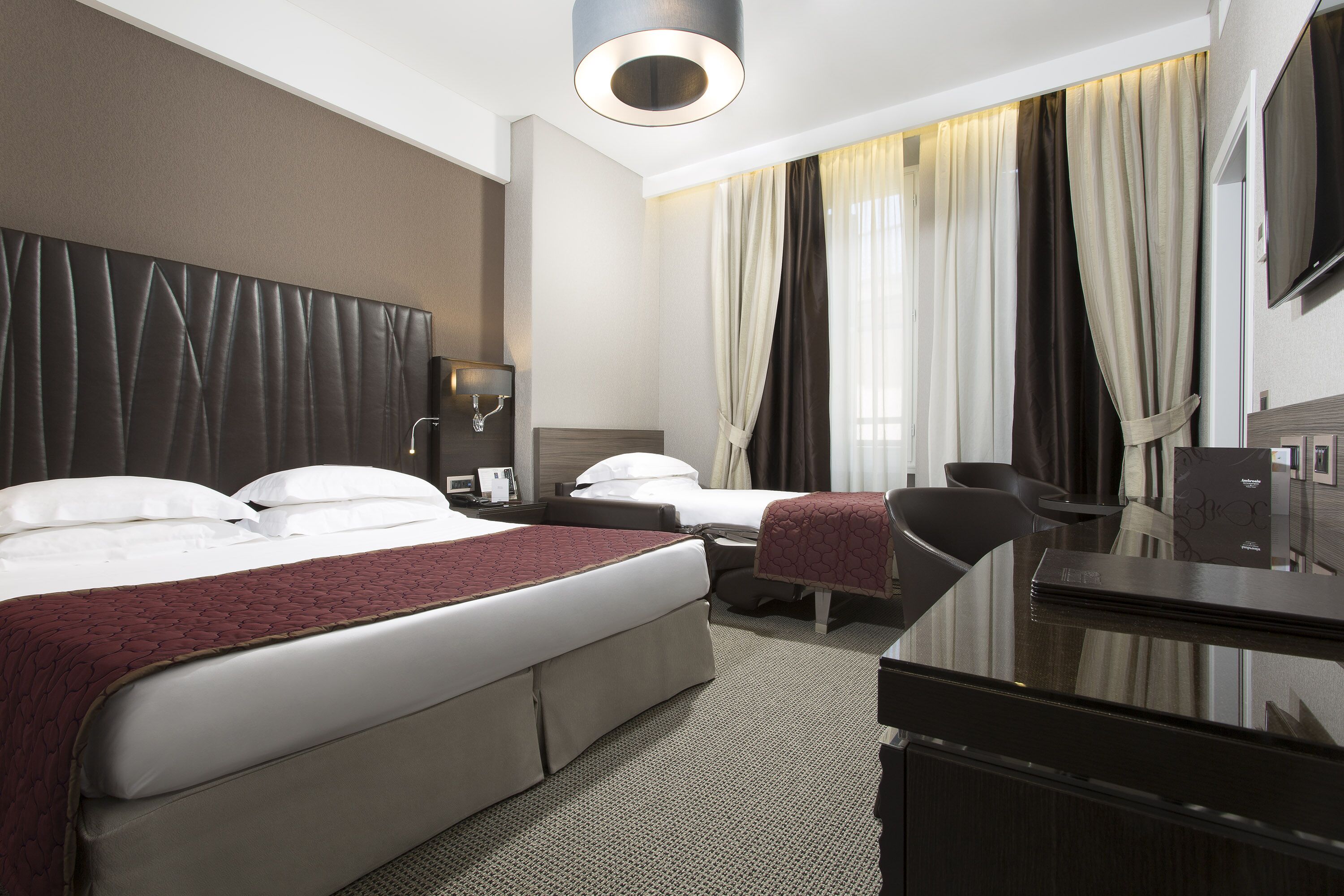A hotel room with two beds, a desk, a chair, a TV, and a large window with curtains.