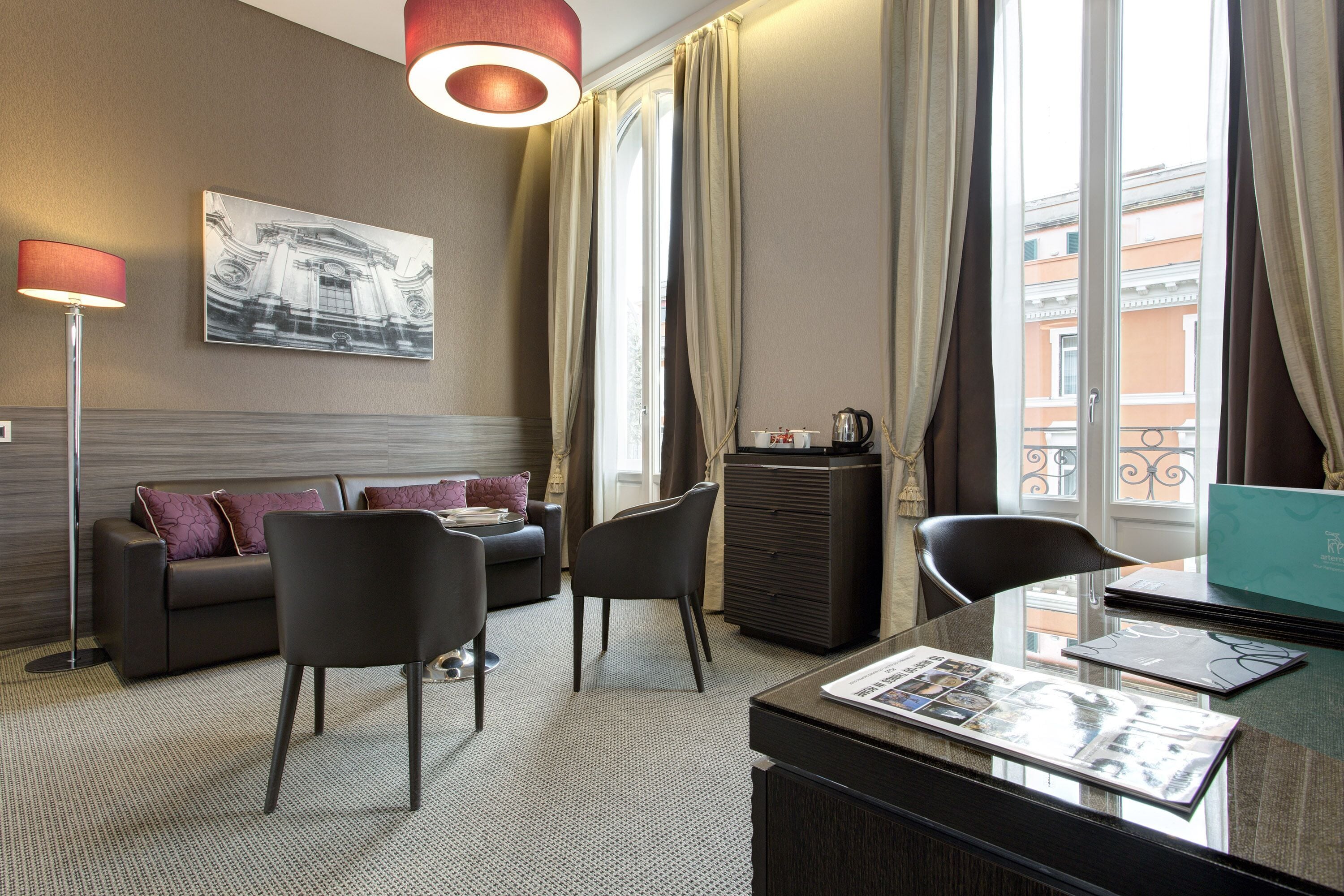 Une chambre d’hôtel avec un canapé, deux fauteuils, un bureau avec un ordinateur portable, une machine à café et un tableau encadré au mur.