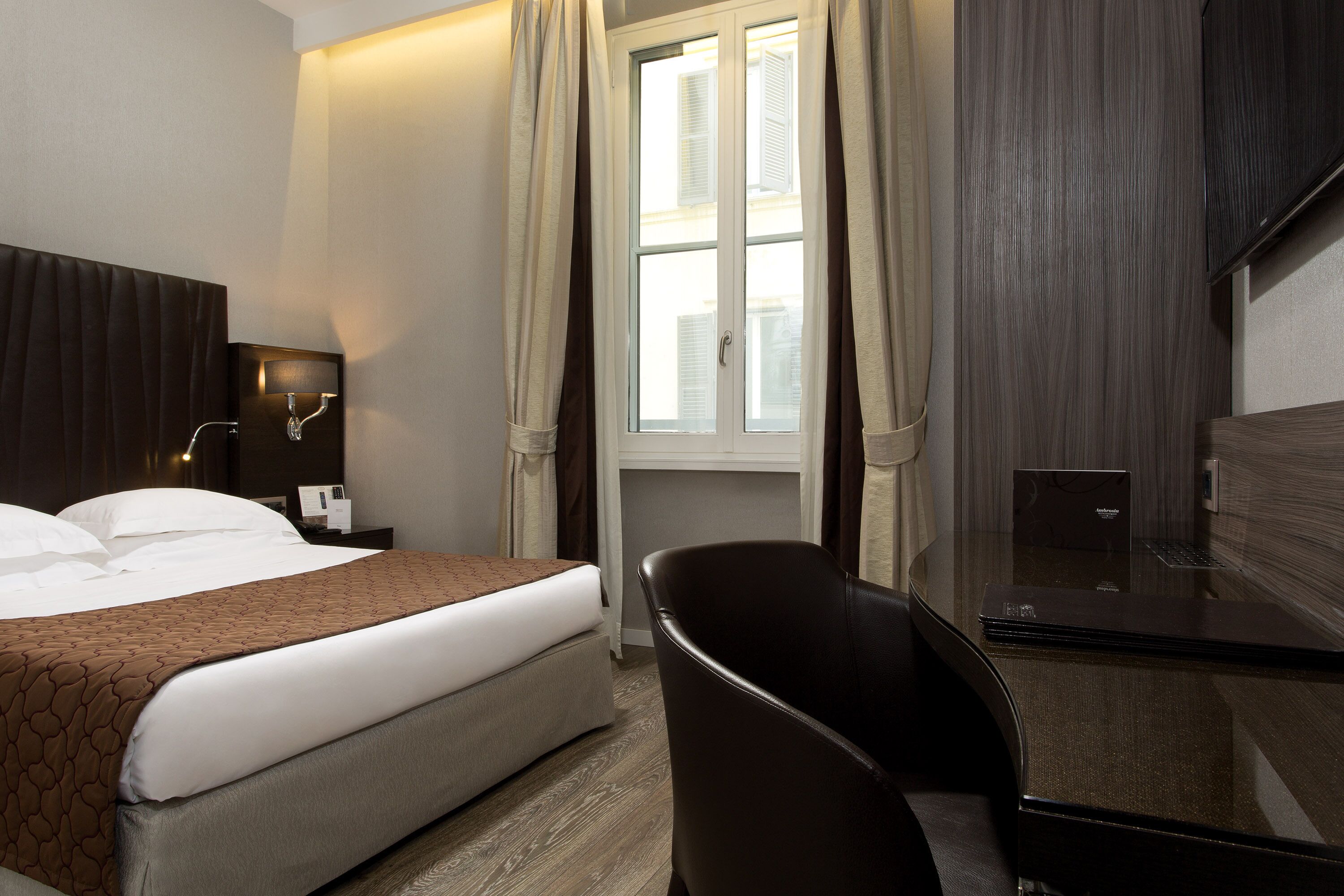 A hotel room with a bed, a desk, a chair, a window with curtains, and a flat-screen TV.