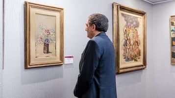 Een man in een blauw pak staat in een kunstgalerie met schilderijen aan de wand.