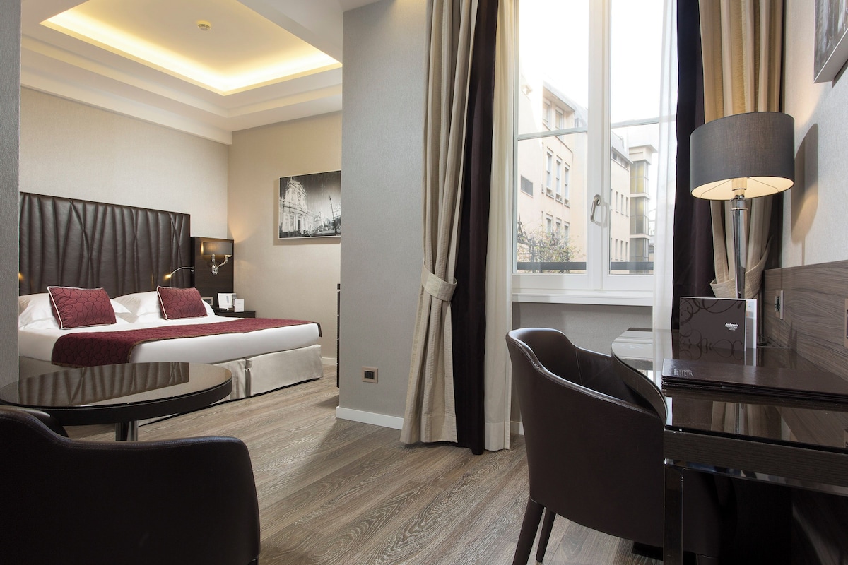 Habitación de hotel con cama, escritorio, silla, lámpara y ventana con cortinas.