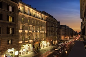 Un edificio histórico de varios pisos, con detalles arquitectónicos ornamentados, y una calle frente a él con automóviles y escaparates iluminados.
