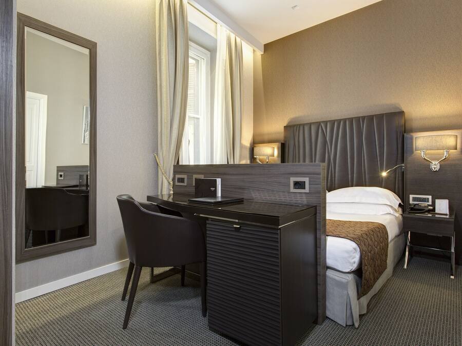 A hotel room with a bed, desk, chair, and a large mirror.