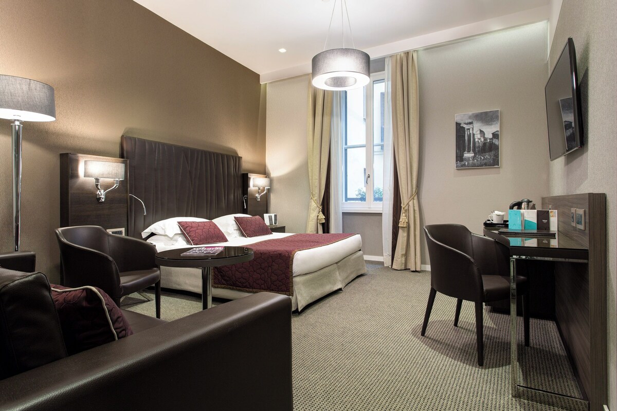 Habitación de hotel con cama, escritorio, silla, lámpara y ventana con cortinas.