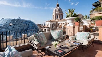 Eine Dachterrasse mit einer Glaskuppel, ein weißes Kuppelgebäude im Hintergrund und eine Terrassenfläche mit einer Couch, Kissen sowie einem Tisch mit Büchern und Blumen.