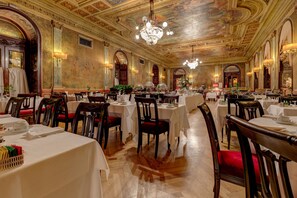A grand dining hall with ornate ceilings, chandeliers, and classical architecture. Tables are set with white tablecloths and red chairs. - Grand Hotel Plaza (Rome)