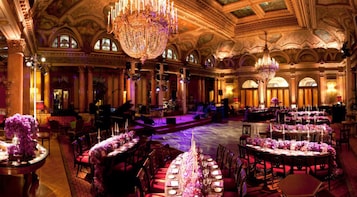 Un salón de baile majestuoso con candelabros elaborados, un escenario y mesas redondas dispuestas para un evento formal.