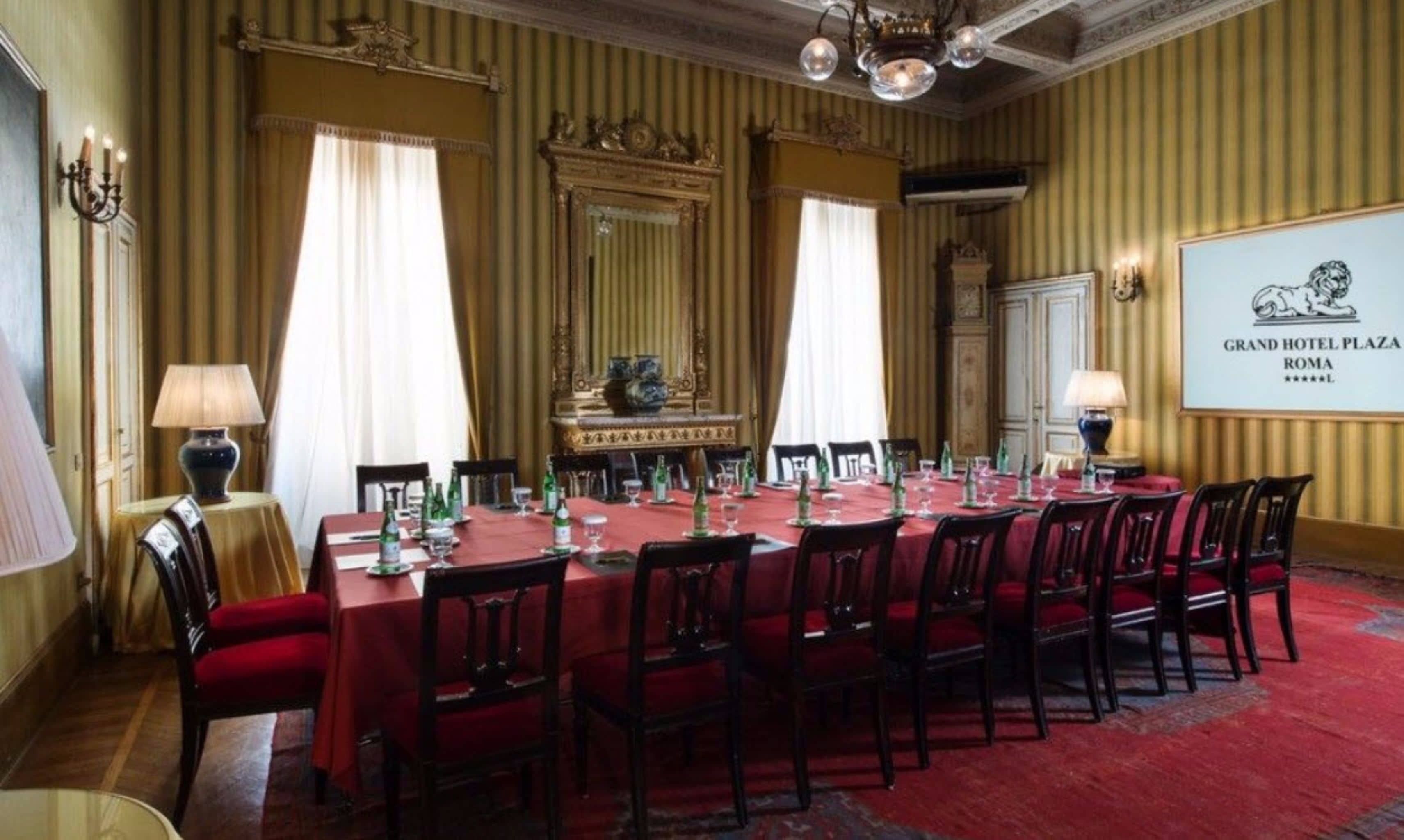 Une salle à manger élégante, avec une longue table dressée pour un repas, des décorations murales raffinées et une enseigne indiquant qu’il s’agit du Grand Hotel Plaza, à Rome.