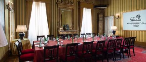 Une salle à manger élégante, avec une longue table dressée pour un repas, des décorations murales raffinées et une enseigne indiquant qu’il s’agit du Grand Hotel Plaza, à Rome.