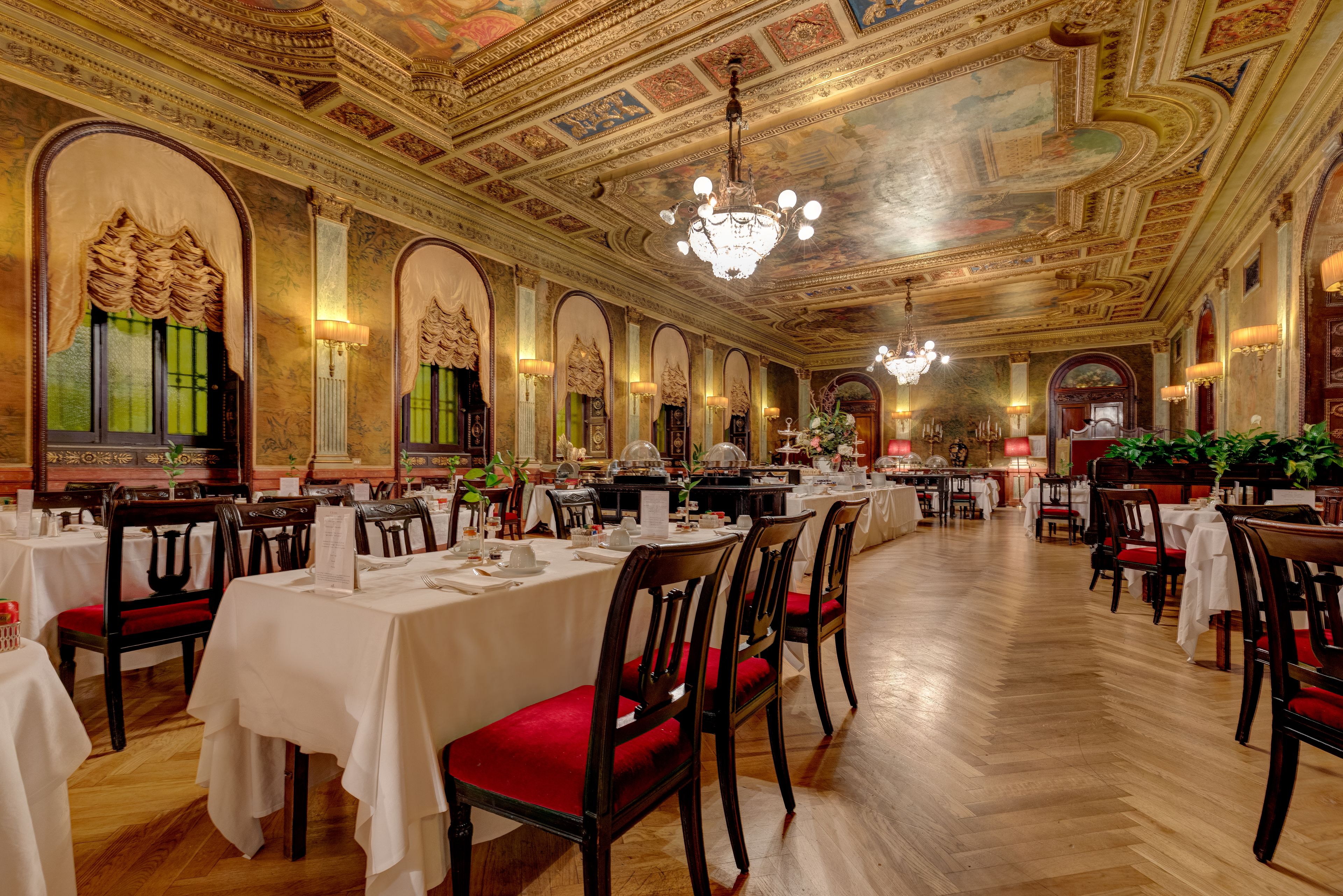 A grand dining hall with ornate ceiling and chandeliers, wooden tables with red upholstered chairs, and a polished wooden floor.
