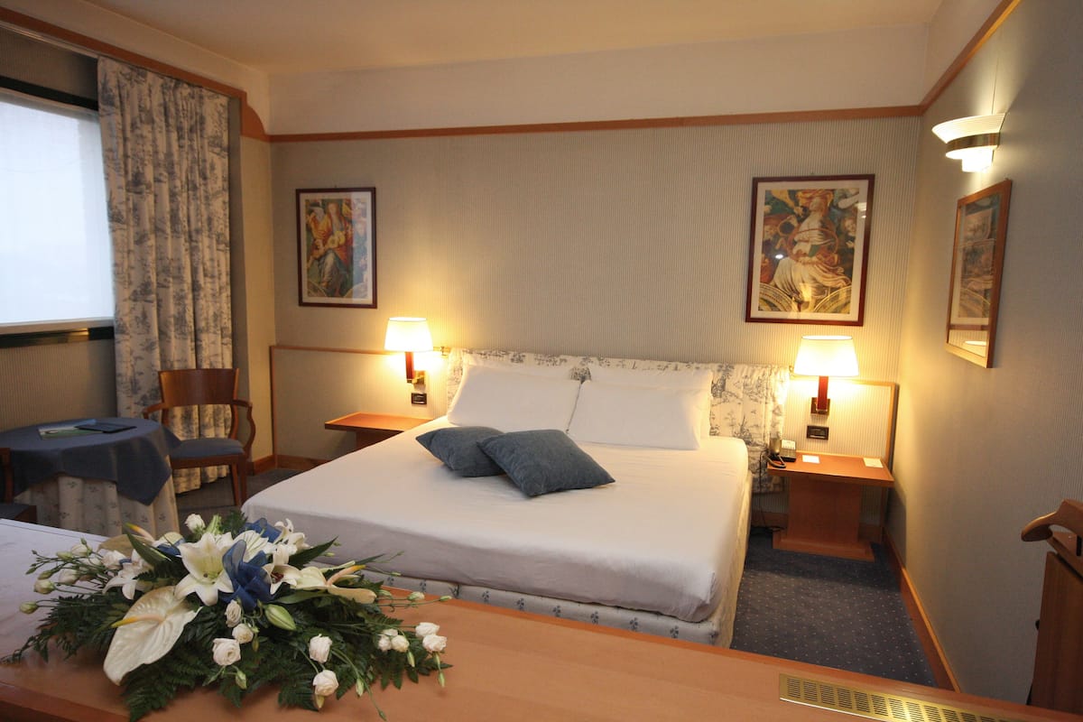 A hotel room with a neatly made bed, two bedside lamps, a desk with a chair, and a floral arrangement on the desk.