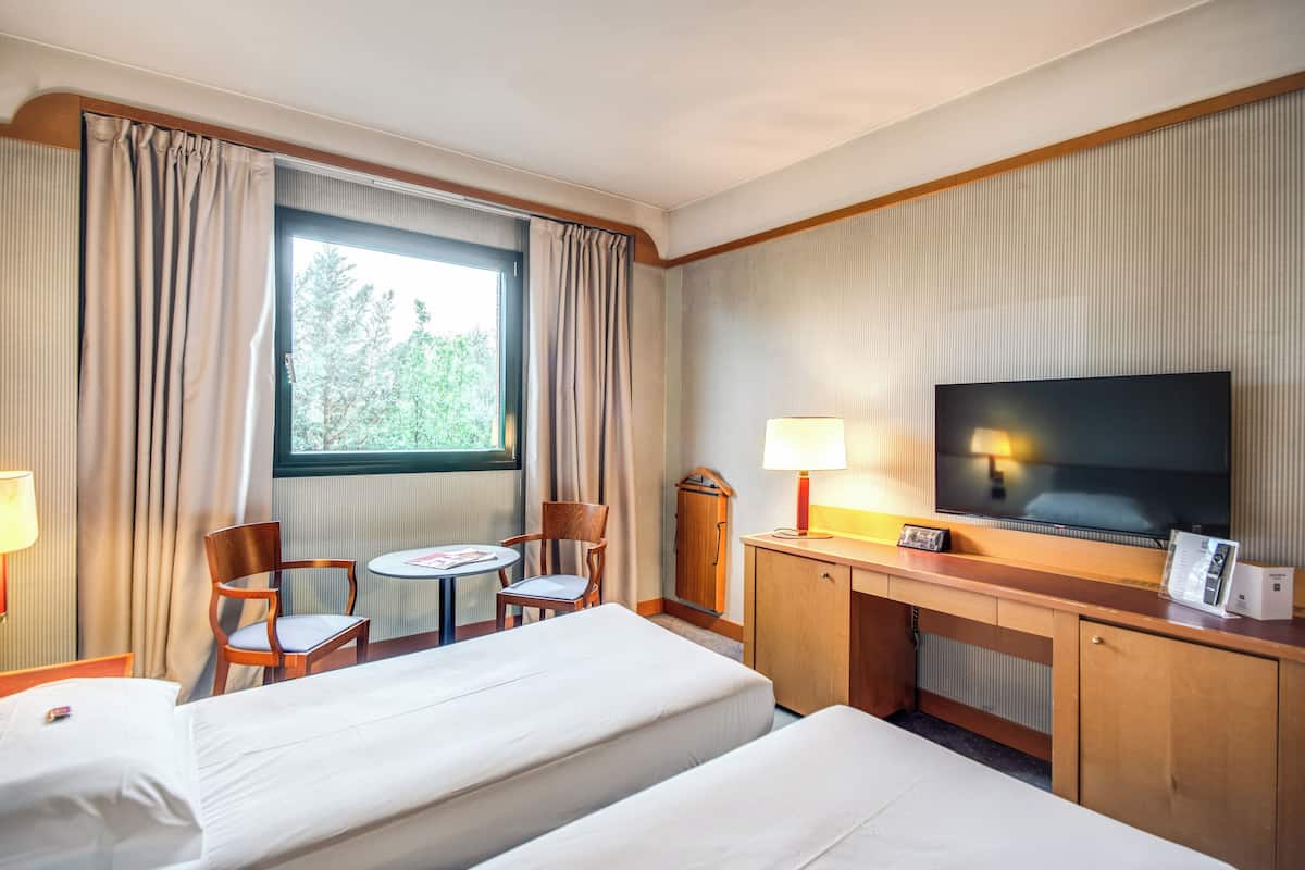 A hotel room with two beds, a desk with a TV, a chair, a small table, and a window with curtains.
