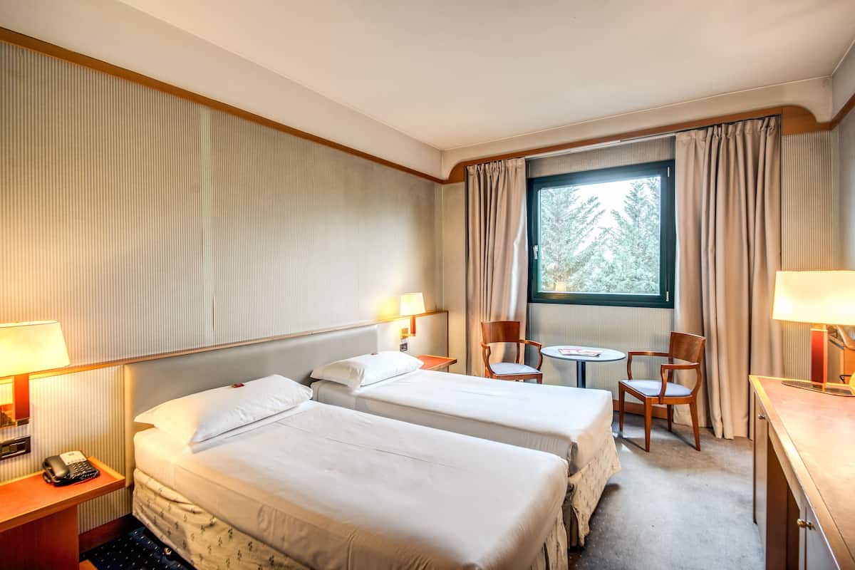 A hotel room with two beds, a small round table, two chairs, a desk with a lamp, and a window with curtains.