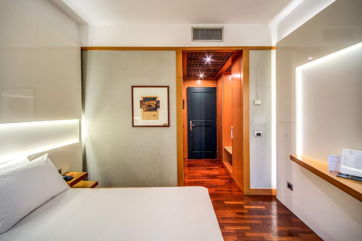 A hotel room with a bed, a nightstand, a framed picture on the wall, a wooden floor, and a door leading to another room.