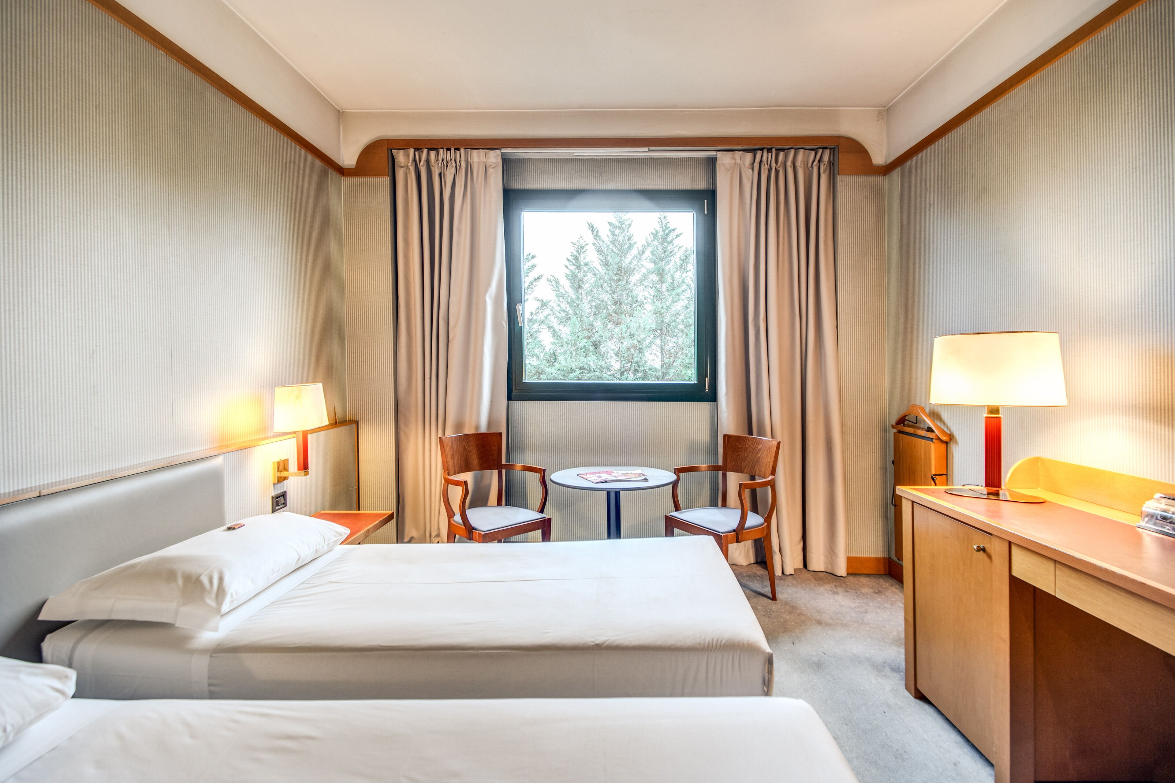 Quarto de hotel com duas camas, uma escrivaninha, uma cadeira, uma mesinha e uma janela com cortinas.