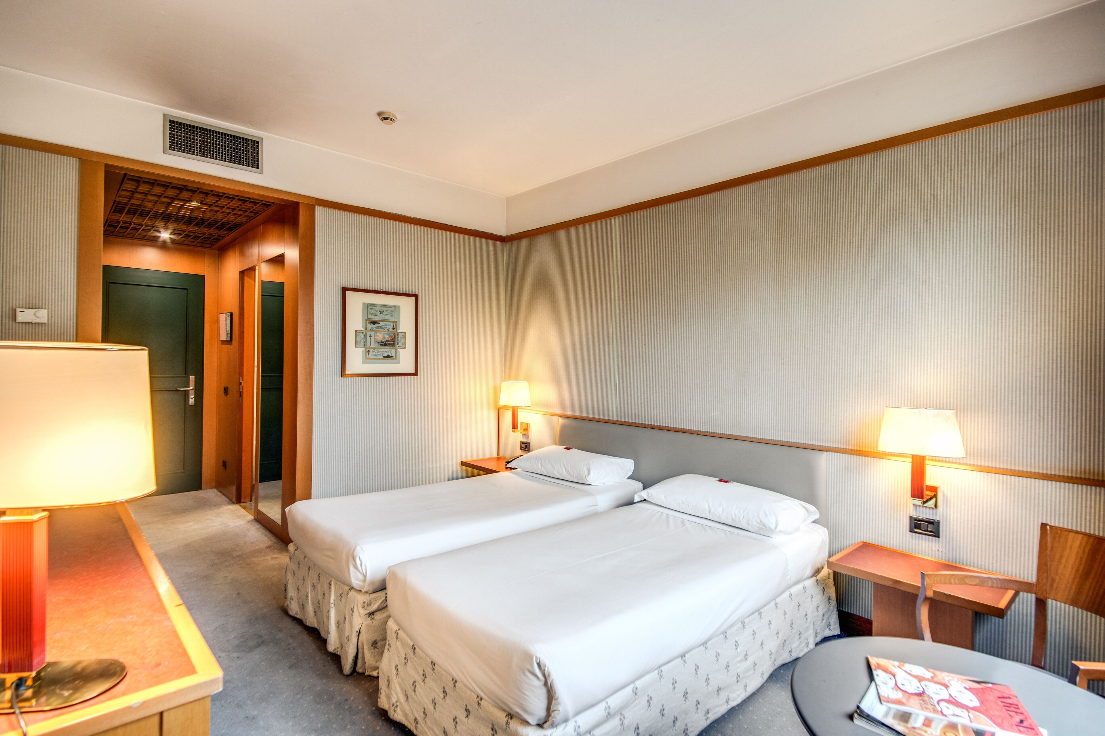 Quarto de hotel com duas camas, uma escrivaninha, uma cadeira, um abajur e uma revista no chão.