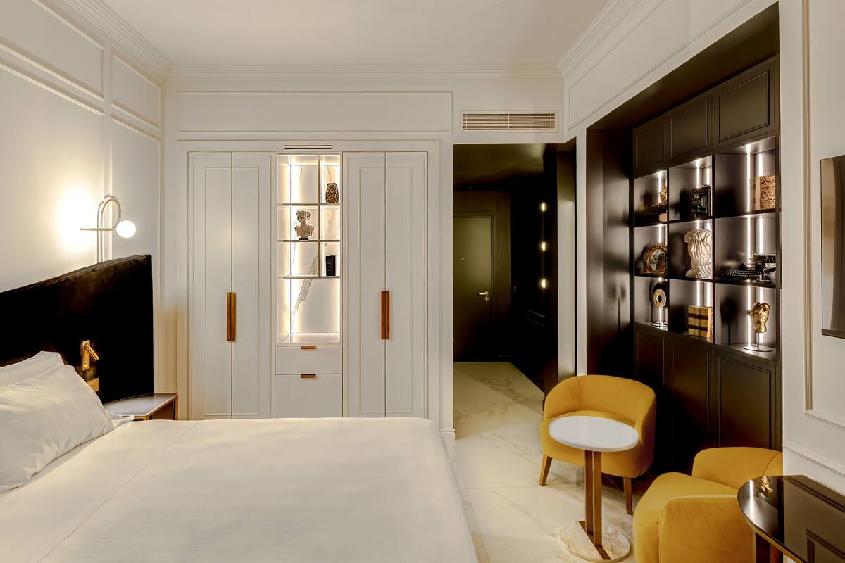 A bedroom with a large bed, bedside tables, a wardrobe, a chair, and a shelf with decorative items.