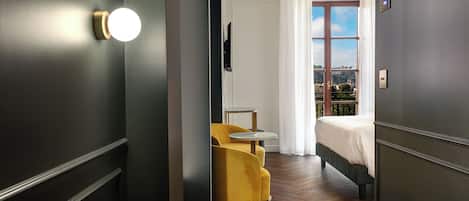 A modern hotel room with a dark wood floor, a bed with a white headboard, a yellow chair, and a large window with a view.