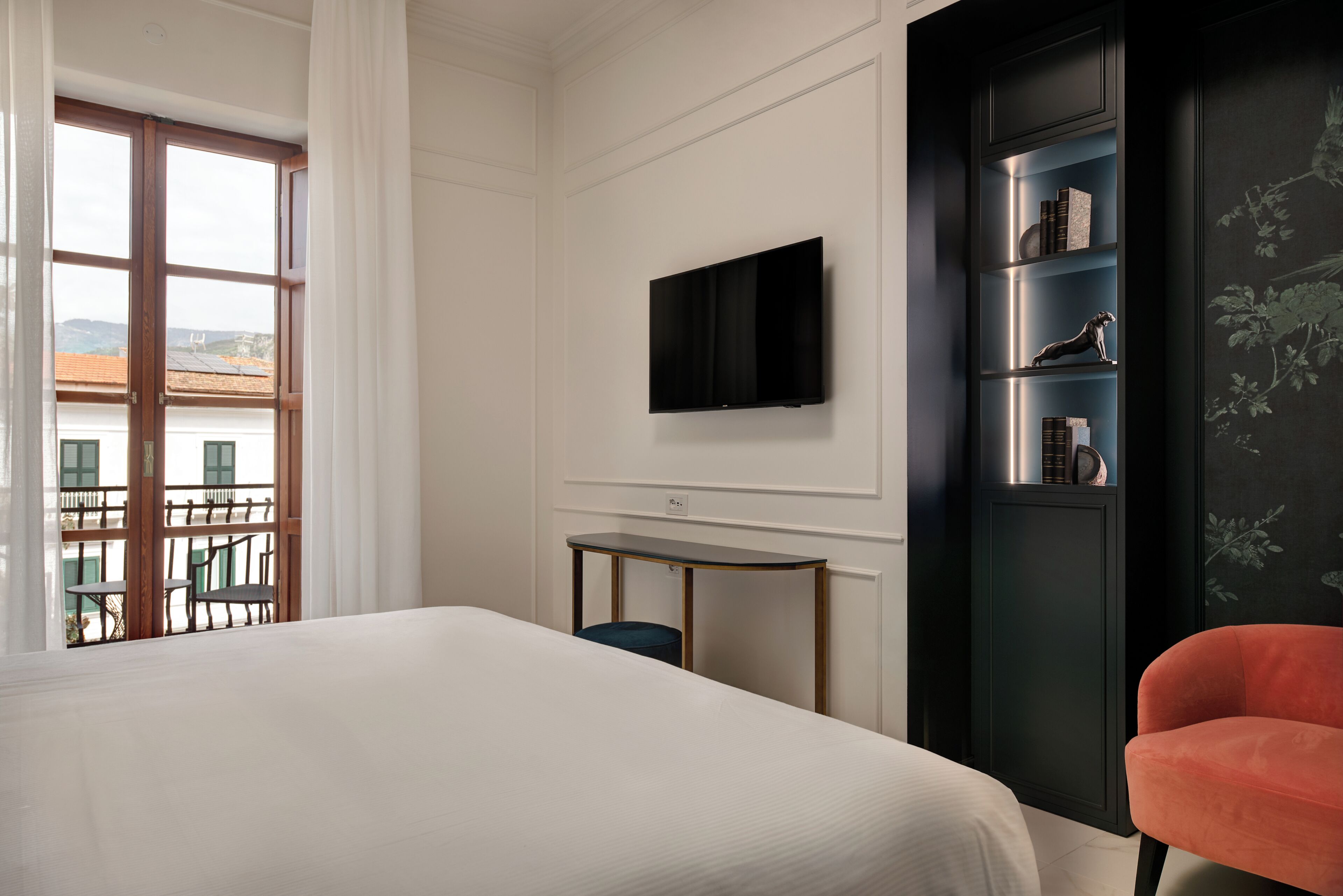 A hotel room with a bed, a TV mounted on the wall, a small table, a chair, and a view of the outside.