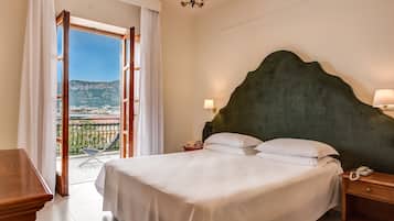 A hotel room with a large bed, a wooden nightstand, a bedside lamp, and a view of the sea through an open door.