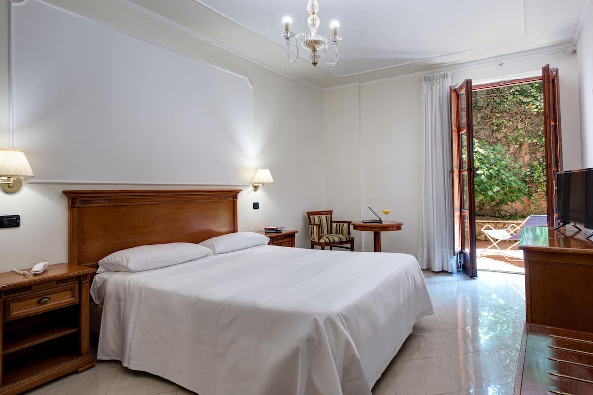 A hotel room with a large bed, bedside tables, a desk with a chair, a TV, and a view of the outdoors.