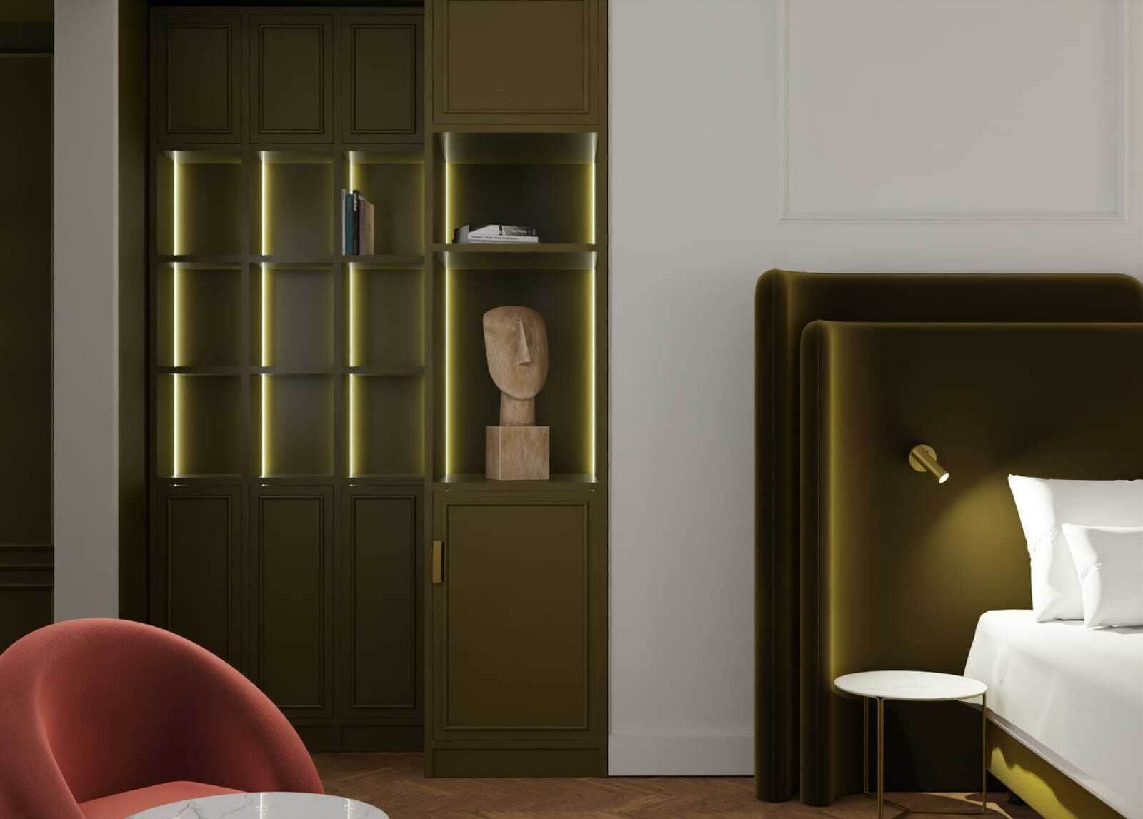 A hotel room with a bed, a red chair, a small round table, and a green shelving unit with books and a sculpture.