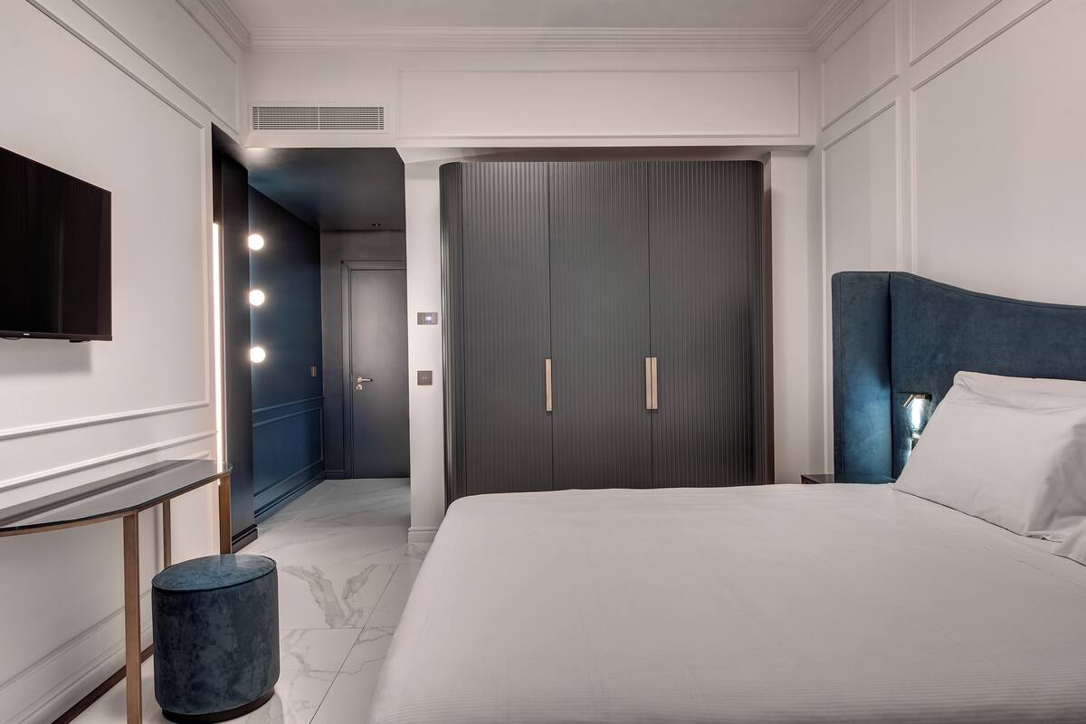 A modern hotel room with a large bed, a television, a vanity with lights, and a walk-in closet.