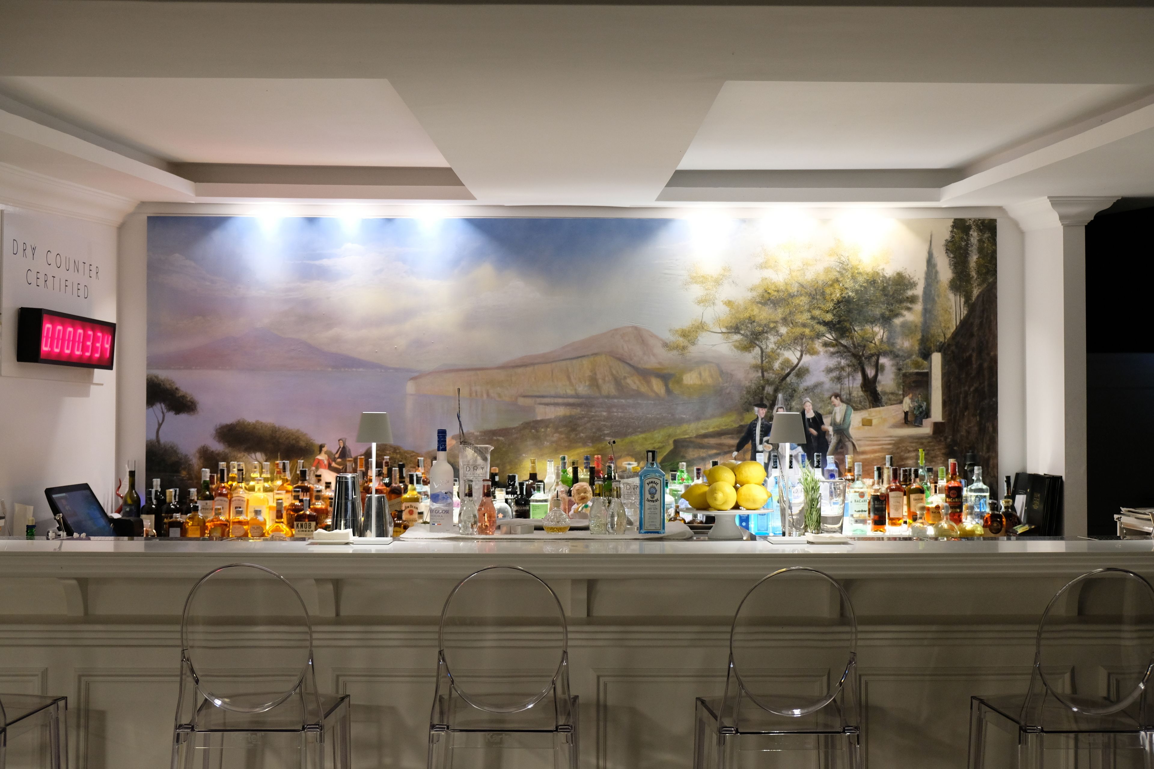 A bar area with a mural, bar stools, and a variety of bottles.