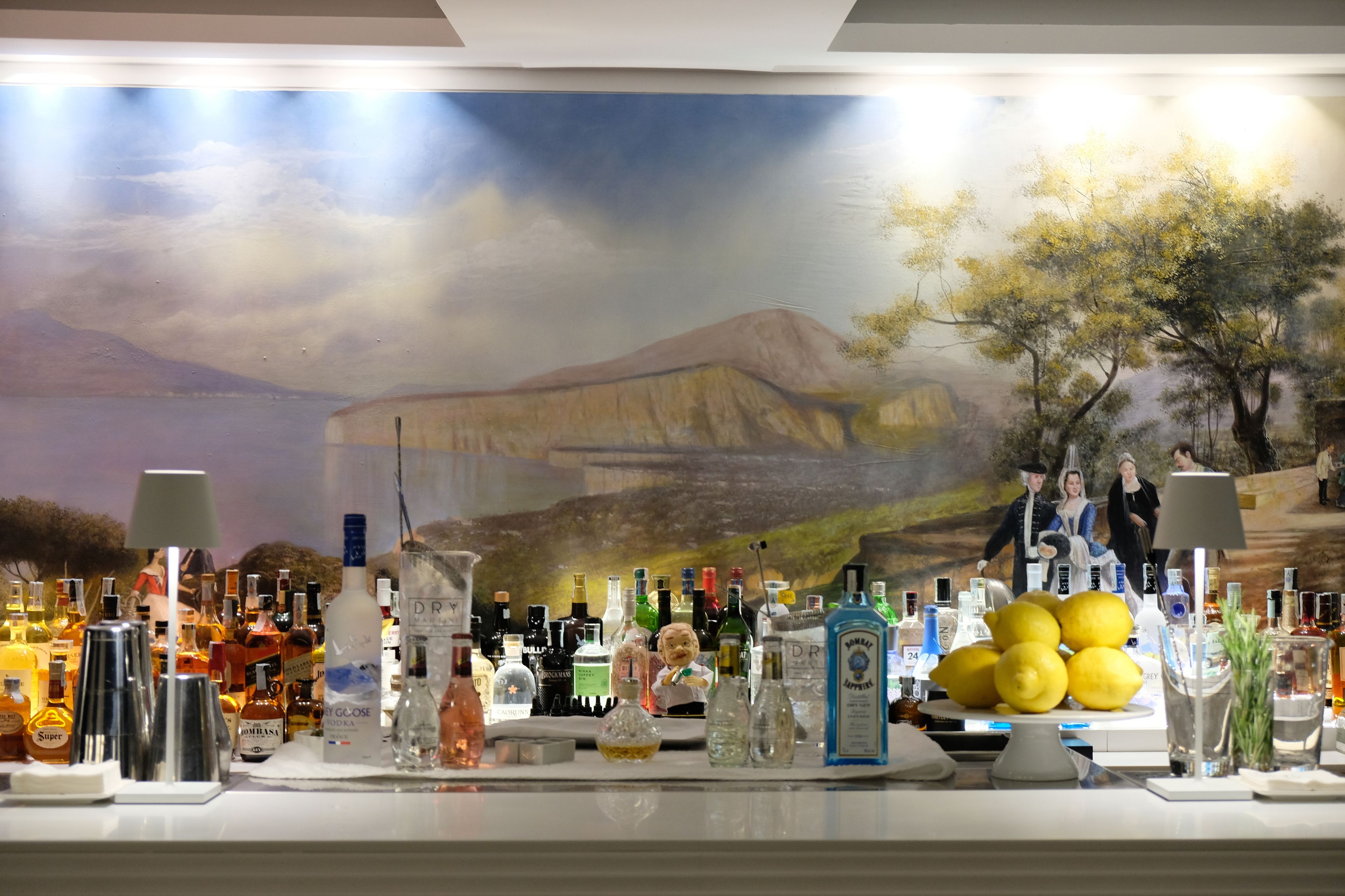Zona de bar con un mural de un paisaje y una variedad de botellas de licor y vasos.
